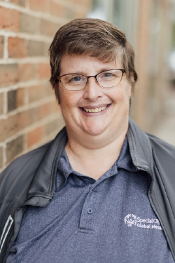 Headshot of Kayte Barton smiling, with short brown hair, wearing glasses and a polo Special Olympics shirt with zip up jacket over top. 