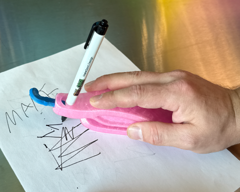 An assistive device that looks like a large ergonomic computer mouse grips a pen and is being moved around with someone's hand