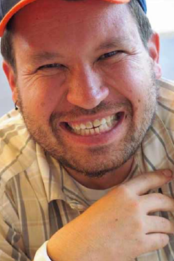 Headshot of Micah Fialka Feldman smiling, wearing a baseball cap and white and gray striped button-up shirt. 