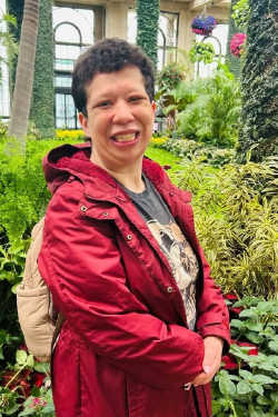 Headshot of Shawna Dedonado smiling, with short dark hair, wearing a backpack and a red jacket over a dark t-shirt.