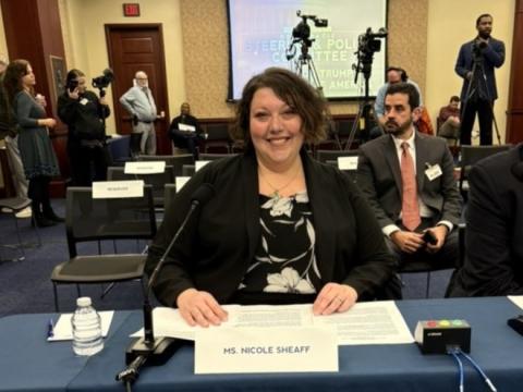 Photograph of a formal hearing with a woman seated at a table speaking into a microphone. Background shows other attendees, media personnel with cameras.
