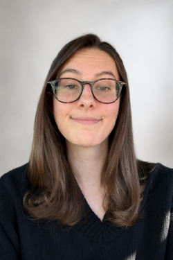 Headshot of Maddie Brodeur smiling, with long brown hair, wearing glasses and a black sweater. 