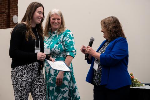 Photograph showing three women engaged in a presentation of award ceremony, with one woman holding a microphone and another holding a certificate.
