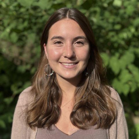 Victoria Petricca is a young woman with wavy brown hair, wearing hoop earrings, and smiling, pictured outside in front of some bushes.