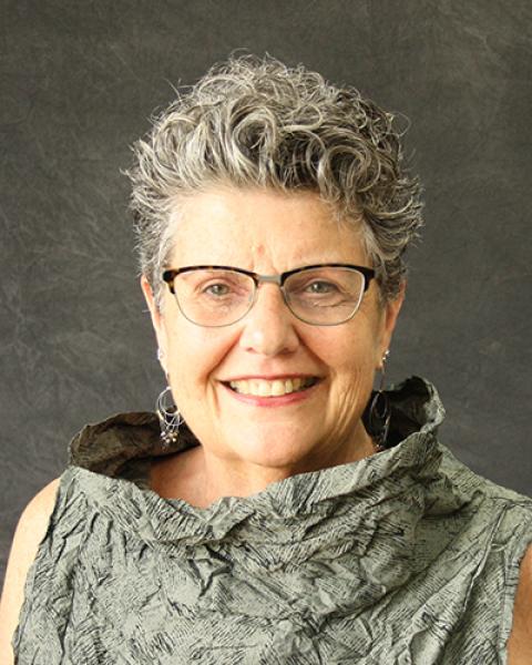 Joan Beasley is a woman with very short curly styled grey hair wearing glasses with a dark frame at the top and silver dangly earrings.