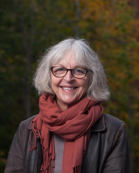A middle-aged woman with shoulder-length gray hair, wearing glasses, a leather jacket, and an orange scarf.