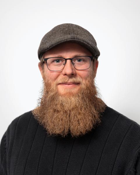 Nate is a man with glasses, and a long red-brown beard, wearing a cap and black shirt.