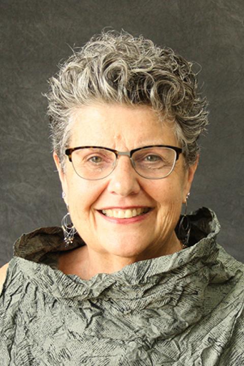 Joan Beasley is a woman with very short curly styled grey hair wearing glasses with a dark frame at the top and silver dangly earrings.
