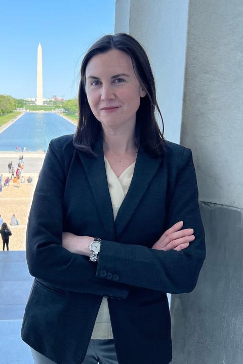 Megan Henly is a woman with shoulder length brown hair in a dark blue blazer leaning up against a stone wall with the Washington Monument and Reflecting Pool in the background