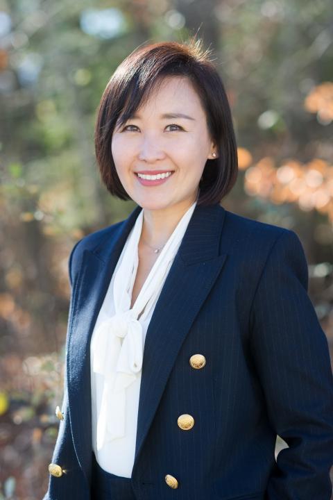 Hyun Ju Kim is a woman with straight brown hair styled in a bob. Shes wearing a white blouse with a navy blue blazer with big gold buttons.