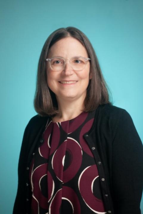 A middle-aged woman with short brown hair and glasses. She wears a shirt with maroon, white, and black swirls and a black cardigan.