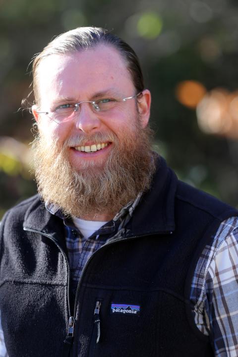 Nathan Thomas is a man with long hair pulled back, a long reddish blond beard, who is wearing glasses, a blue plaid flannel with a fleece vest over it 