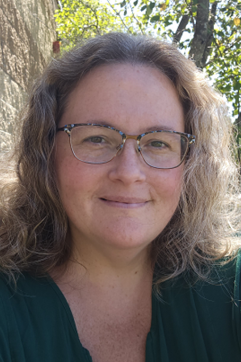 Tina Kelleher is a woman with wavy shoulder-length brown and grey hair. She's wearing a green blouse and has on glasses with green and blue specks on the top half of the frame.