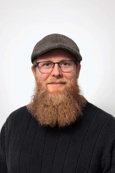 Nathan is a man with glasses, and a long red-brown beard, wearing a cap and black shirt.