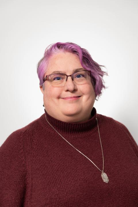 Toni Sumner-Beebe is a woman with short purple-pink hair wearing glasses and a maroon shirt.