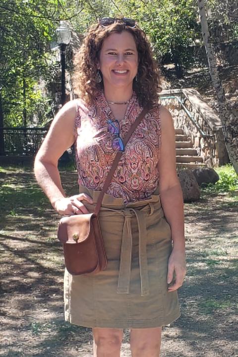 Tracey Parent is a woman with curly shoulder length brown hair. She is smiling and posing outside in a park on a sunny day.