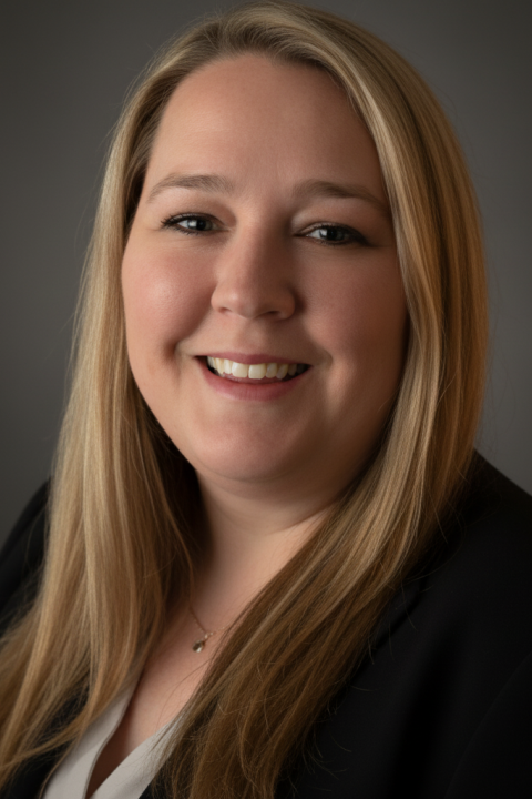 Tiffany Liska is a woman with straight blonde hair, wearing a black blazer and necklace, standing in front of a grey background.