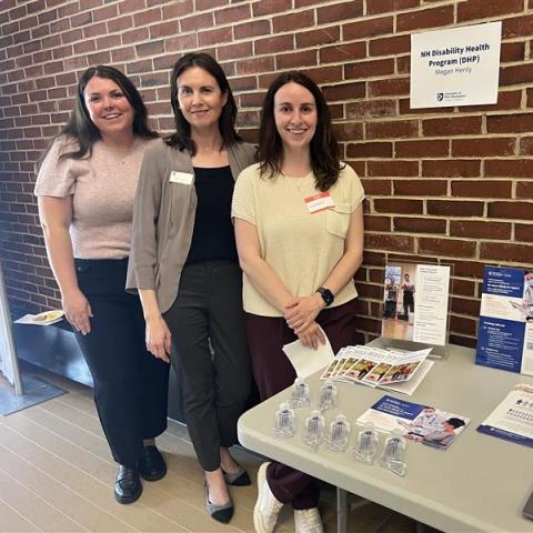     Three women pose next to a sign that reads “NH Disability and Health Program (DHP) and a table with promotional material.  
